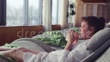 成熟的女人，穿着白色的晨衣，躺在水疗中心，躺在沙发上喝茶，看着窗外。
